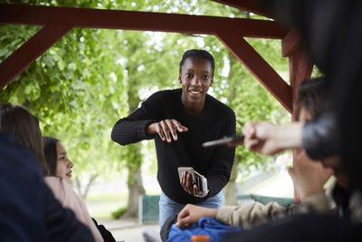 Smiling teenage girl collecting smart phone from friends while enjoying at park