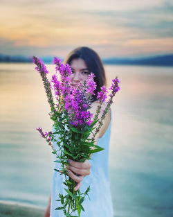 Portrait of woman holding pink flowers while standing against sea and sky