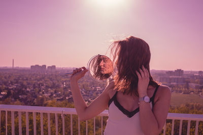 Woman holding mirror in city against clear sky