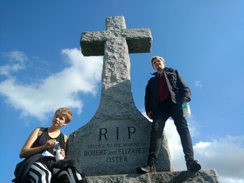 Low angle view of young woman and boy by cross at cemetery against sky