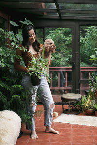 Full length of young woman holding potted plant and carrying poodle while standing on porch
