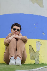 A girl in dark glasses sits near a wall 