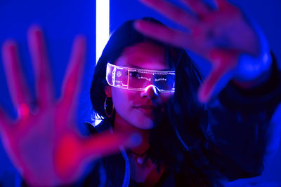 Ethnic female touching air while experiencing virtual reality in modern transparent vr glasses in dark studio with bright neon illumination looking at camera