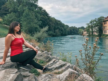 Young woman sitting on rock by lake against sky
