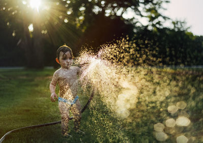 Shirtless baby girl spraying water with garden hose at yard