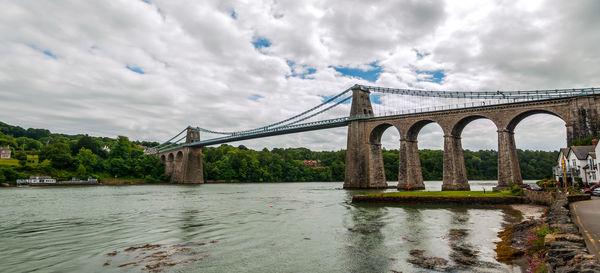 Menai bridge over river against cloudy sky