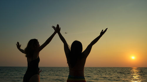 Silhouette friends with arms raised against sea during sunset