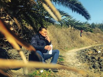 Young man sitting on tree against sky