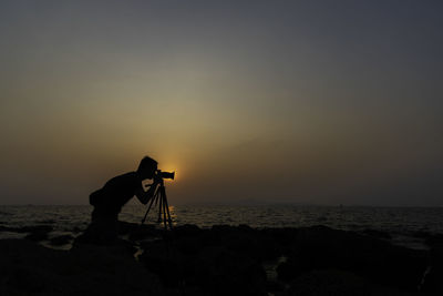 Silhouette man photographing sea against sky during sunset