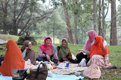 Smiling women wearing hijabs while sitting on picnic blanket in forest