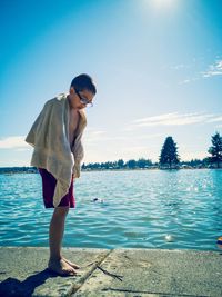 Full length of wet boy with towel standing by lake against blue sky during sunny day