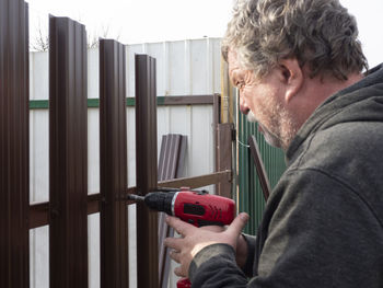Side view of senior man drilling metallic fence