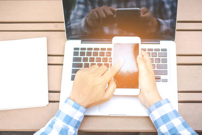 Cropped hands of man using mobile phone with laptop at table