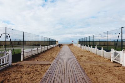 Boardwalk amidst playing field at beach against cloudy sky