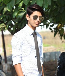 Portrait of young man wearing sunglasses standing against tree