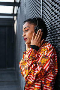 Side view of young thoughtful ethnic woman in colorful trendy shirt touching wireless headphones and listening to music looking away while leaning on gray wall on city street