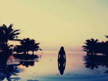 Silhouette woman relaxing in infinity pool amidst palm trees against sea during sunset