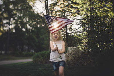 Cheerful girl with american flag running in yard during sunny day