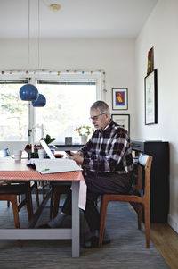 Side view of retired senior man using smart phone while sitting with financial bills at dining table