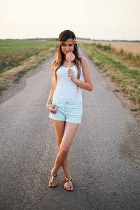 Portrait of smiling young woman holding lollipop while standing on road during sunset