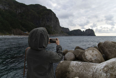 Rear view of woman photographing on rock by sea against sky