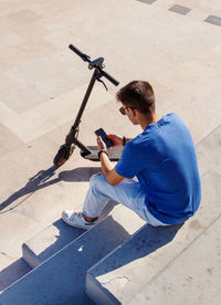 Young caucasian man with smartphone sitting outdoor near electric scooter and using smartphone