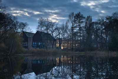 Bare trees by lake and buildings against sky