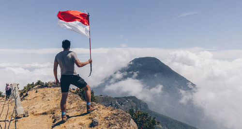 Rear view of man holding flag while standing on mountain against sky