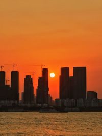 Silhouette buildings by sea against orange sky