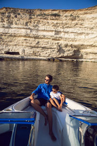 Man with a beard and a boy with son in sunglasses are sitting on yacht near rocks in  sea in summer