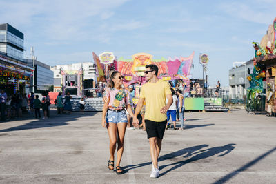 Happy young man in casual clothes and sunglasses holding hand of cheerful trendy female looking at each other strolling in city fairground