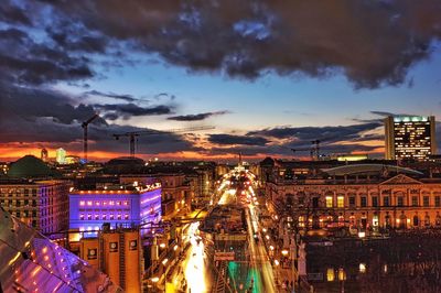 Illuminated street amidst buildings against cloudy sky at dusk in city