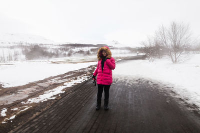 Woman holding camera while standing on footpath amidst snow covered field during winter