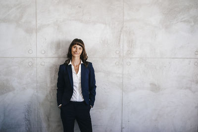 Businesswoman leaning against office wall with hands in pockets