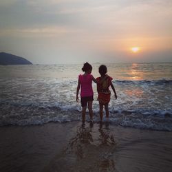 Rear view of friends standing on shore at beach against sky during sunset