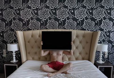 Shirtless man holding television set while sitting on bed at home