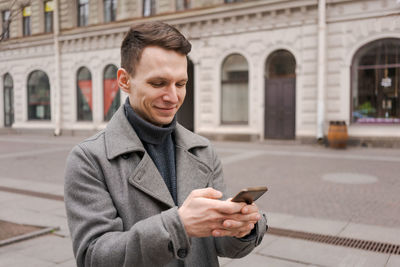 Smiling young man in gray coat is holding scroll text messages in his mobile