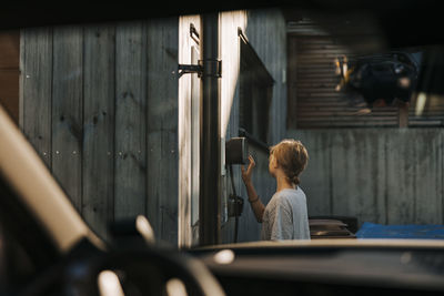 Boy operating car charging device mounted on wall of house seen through windshield