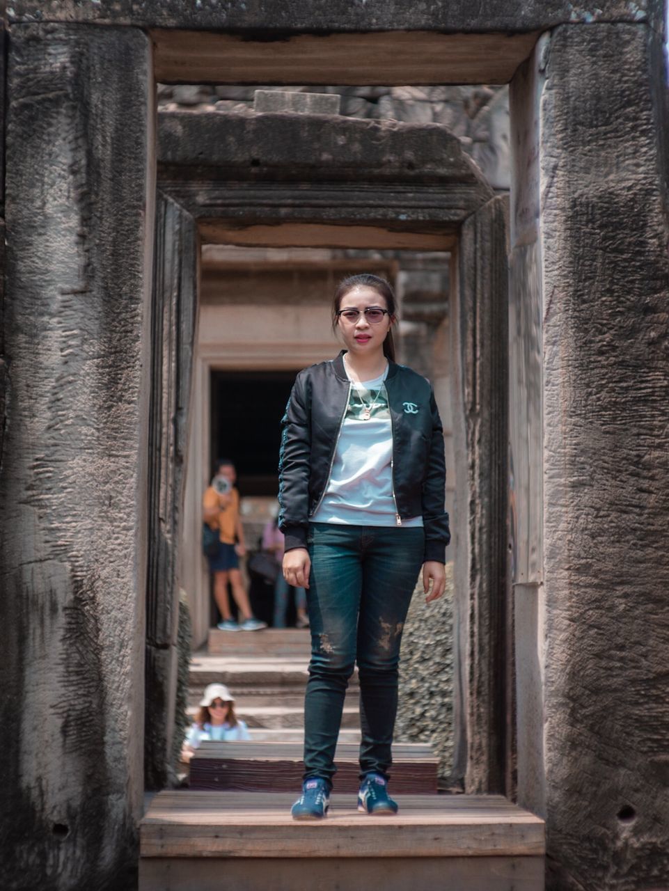 full length, front view, one person, architecture, young adult, looking at camera, portrait, standing, jacket, casual clothing, day, adult, clothing, real people, built structure, hands in pockets, smiling, building exterior, outdoors, architectural column, leather