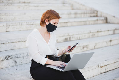 Concentrated businesswoman in elegant clothes and medical mask sitting on stairs on street and working on remote project while using laptop and smartphone