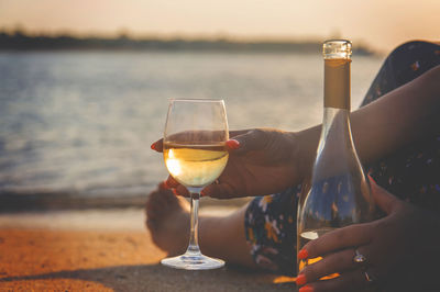 Midsection of man holding beer glass against sky during sunset