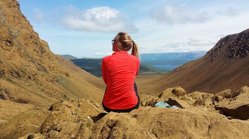 Rear view of woman sitting on rock by mountains against sky