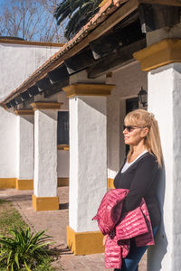 Mature adult blonde woman smiling with glasses with jacket in hand on sunny day.