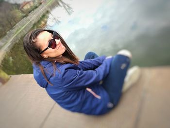 High angle view of smiling young woman in sunglasses sitting on jetty over lake