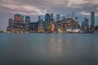 Illuminated city at waterfront against cloudy sky