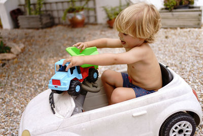 Side view of cute cheerful little boy with blond hair riding toy car while laying in yard on sunny summer day