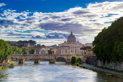 Arch bridge over river by buildings against cloudy sky