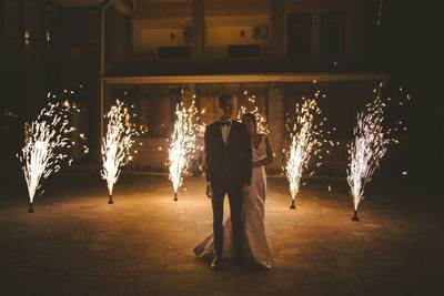 Full length of bride and bridegroom standing by firework display at night