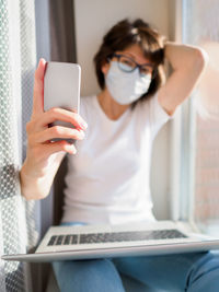 Pretty woman in medical mask remote works from home. she making selfie on window sill with laptop. 