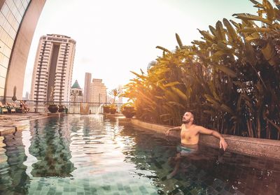 Full length of shirtless man sitting in swimming pool against sky